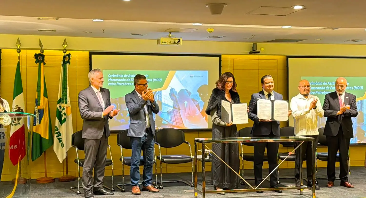 Business leaders at a formal signing ceremony holding agreement documents while attendees applaud on stage