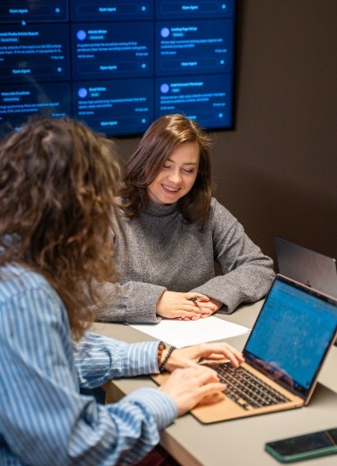 Man and woman standing together at a desk with a laptop and notebooks, smiling at the camera.