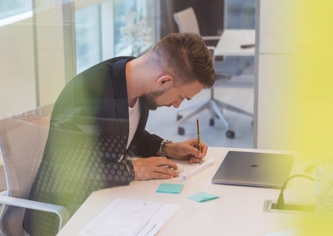 Close-up portrait of a person wearing a gray insulated hooded jacket with the hood up, showing their face framed by the hood against a light background.