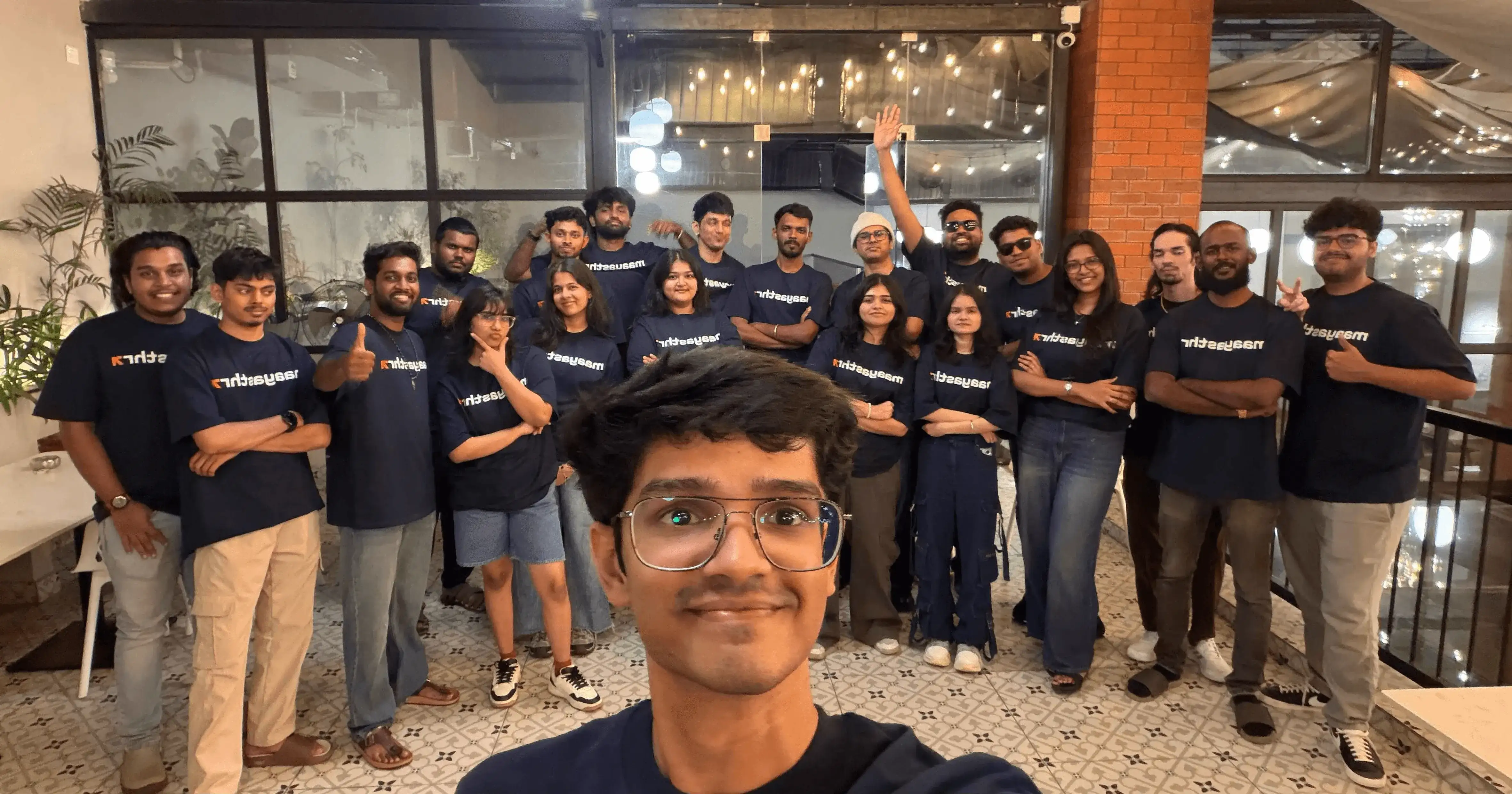 Team selfie featuring Maayasthra members wearing matching branded t-shirts, gathered indoors during a casual team meetup event.