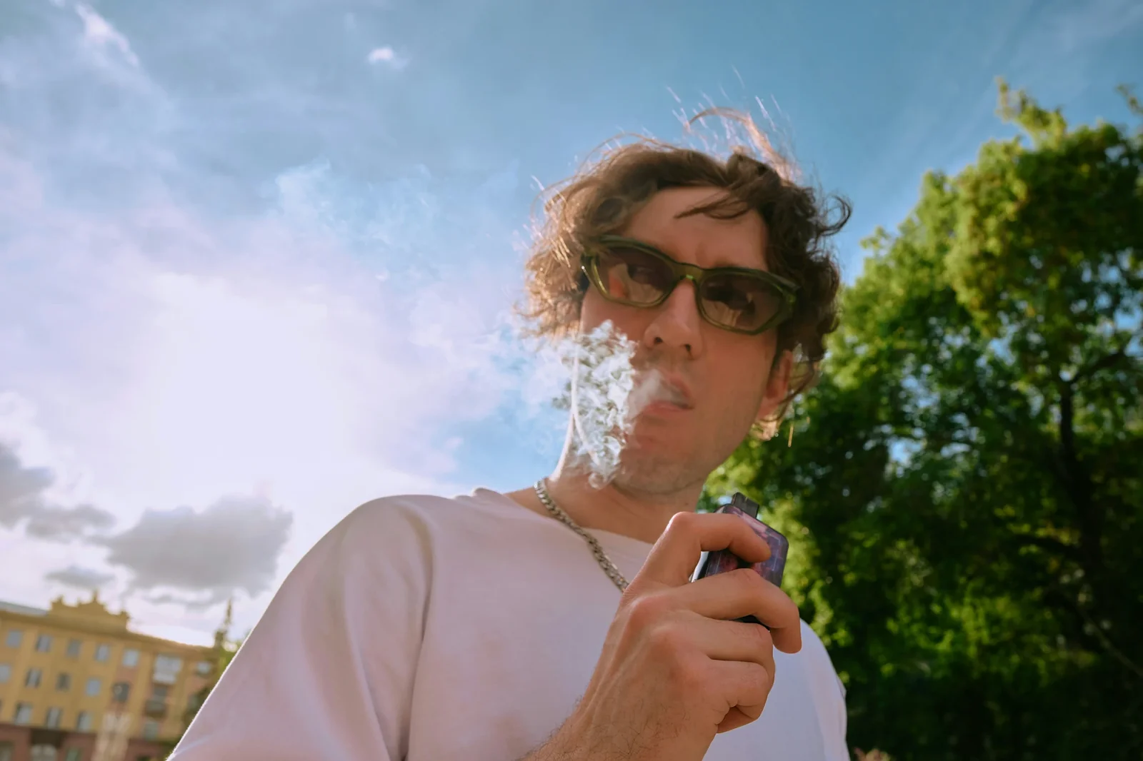 Man in sunglasses exhales vapor outdoors on sunny day with trees and building in background.