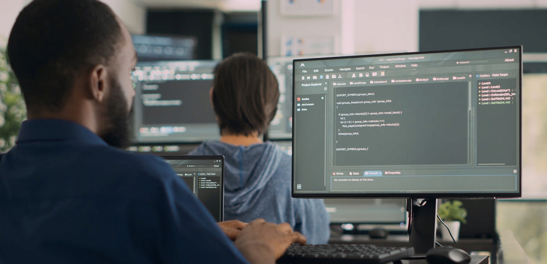 Two individuals working at computer stations, with code displayed on large monitors—representing cybersecurity operations or secure software development in progress.