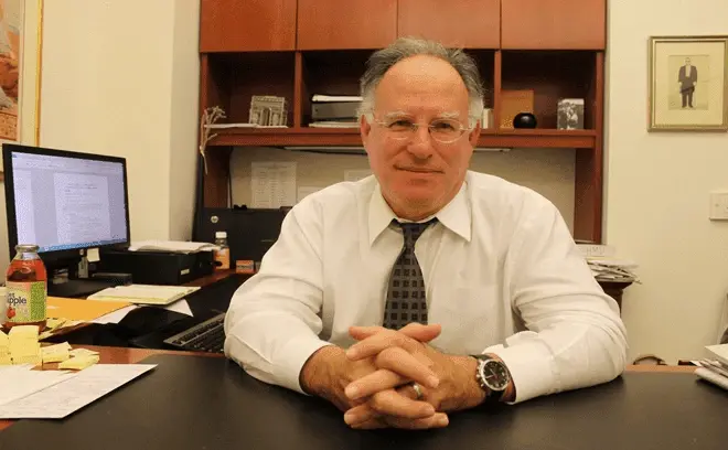 Judge Harry Dorfman's seated at a desk in an office, hands clasped, with shelves and documents behind him.