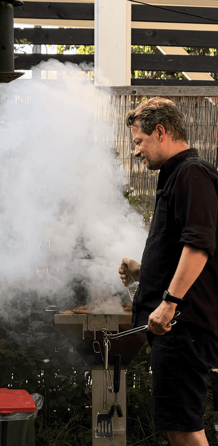 Mann in schwarzer Kleidung steht an einem rauchenden Grill im Garten