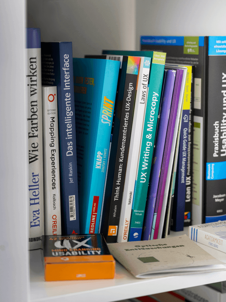 Regal mit Fachbüchern zu UX-Design, Usability und Interface-Themen, darunter Titel wie 'Wie Farben wirken', 'Das intelligente Interface' und 'UX Writing & Microcopy'