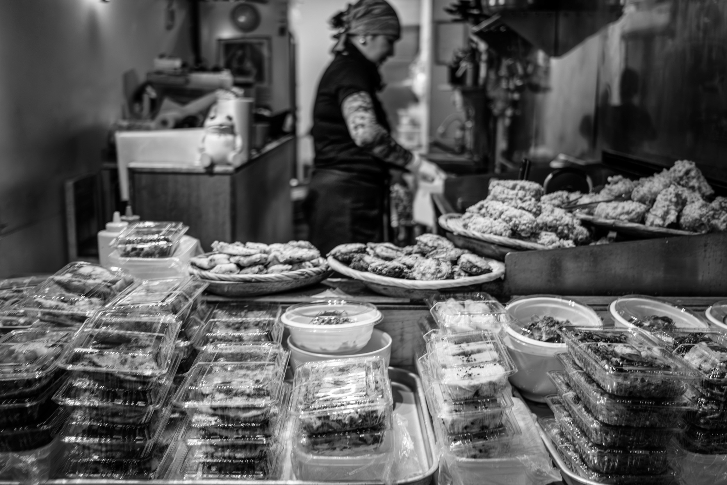 Black and white photograph of trays of prepared fried foods stacked at a small market stall.