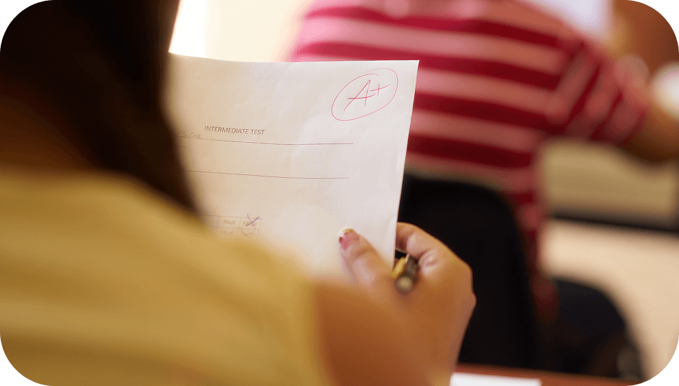 Student holding a returned paper test marked with a red A+ in a classroom.