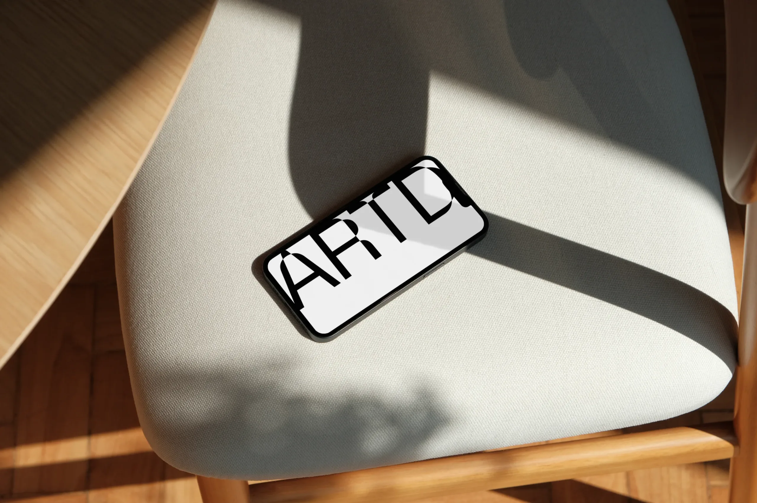 A smartphone mockup placed on a beige upholstered chair with natural light casting soft shadows. The screen displays a purple design with bold typography, creating a stylish contrast with the minimalist wooden furniture.