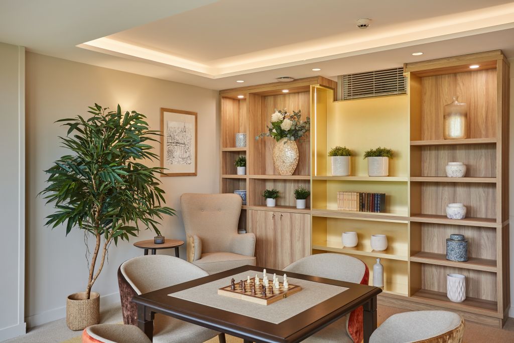 Warm interior living and dining space with wooden shelving, dining table, comfortable seating, potted green plant, and soft ambient lighting