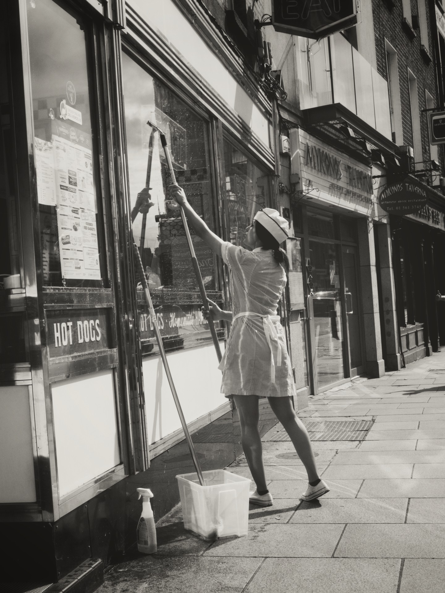 Woman in uniform cleaning restaurant window with bucket and mop on sidewalk beside hot dog shop.