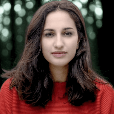 A smiling man with curly hair is sitting at a desk, looking at the camera, with a laptop and a lamp in the background.