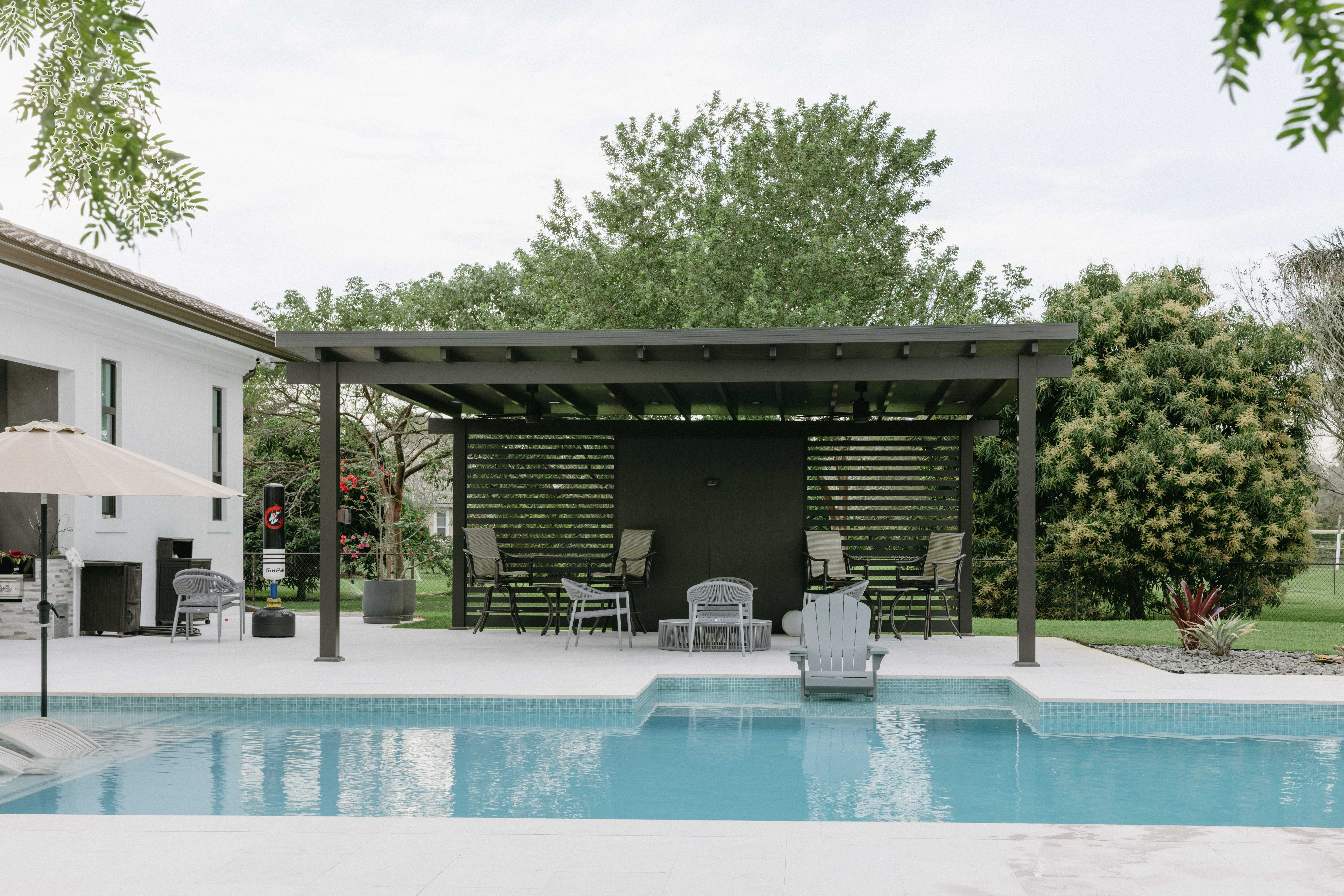 Modern pergola with seating area overlooks a bright blue swimming pool on a sunny day.