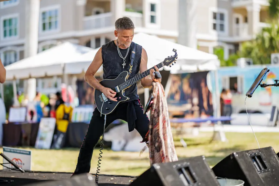 A musician passionately plays electric guitar on stage at a lively outdoor event during BAMFest in Bradenton, FL, with vibrant artwork and festival tents in the background.