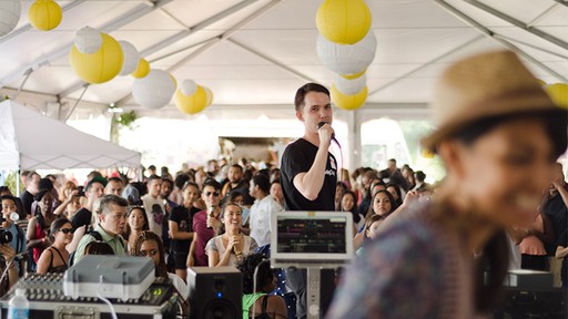 Ken Tanabe on a stage holding a microphone under a festival tent in front of a large audience with a DJ in the foreground at the Loving Day Flagship Celebration in New York City.