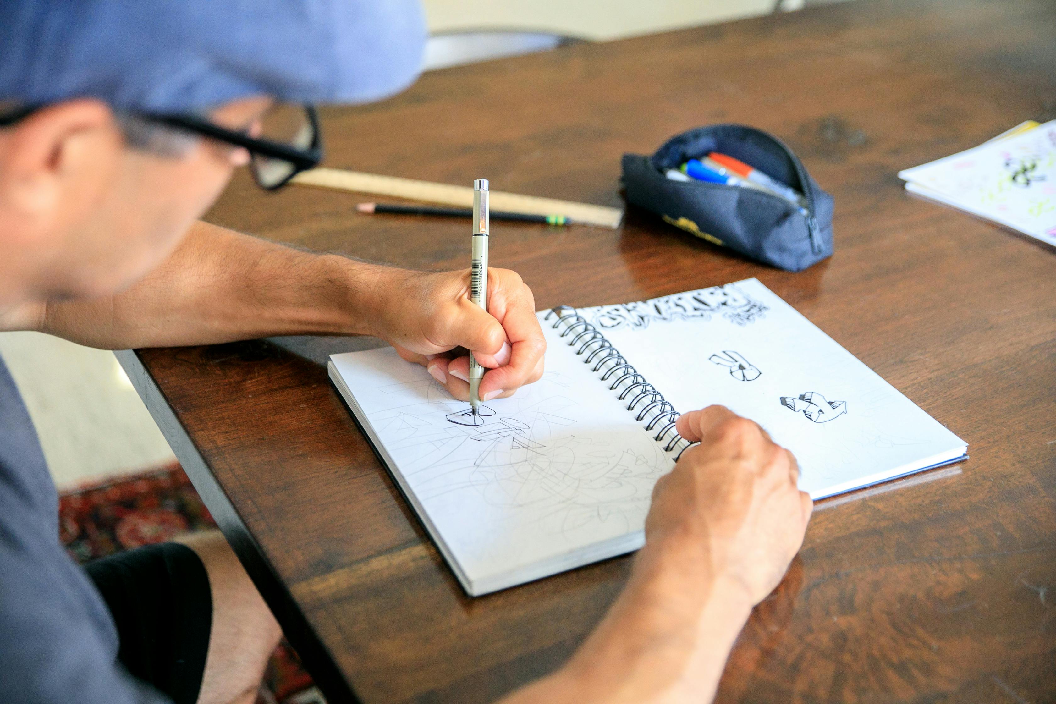 a-man-wearing-glasses-sketches-in-a-notebook-at-a-wooden-table-with-art-supplies - rdne-stock-project (pexels)