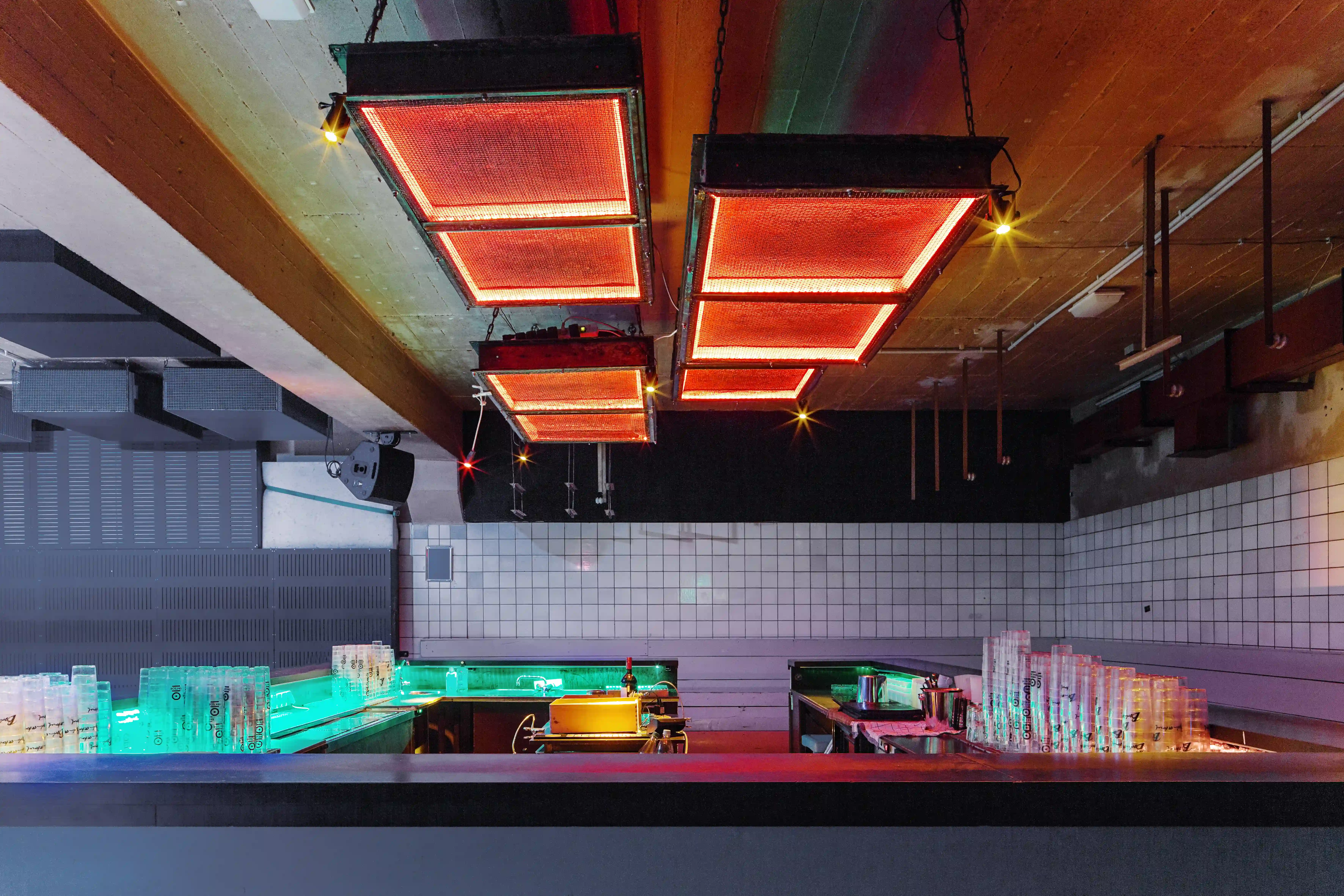 Bar at OHM CLUB with red-glowing heating grids suspended from the raw concrete ceiling, white tiled walls, a dark counter and green backlit bottle displays.