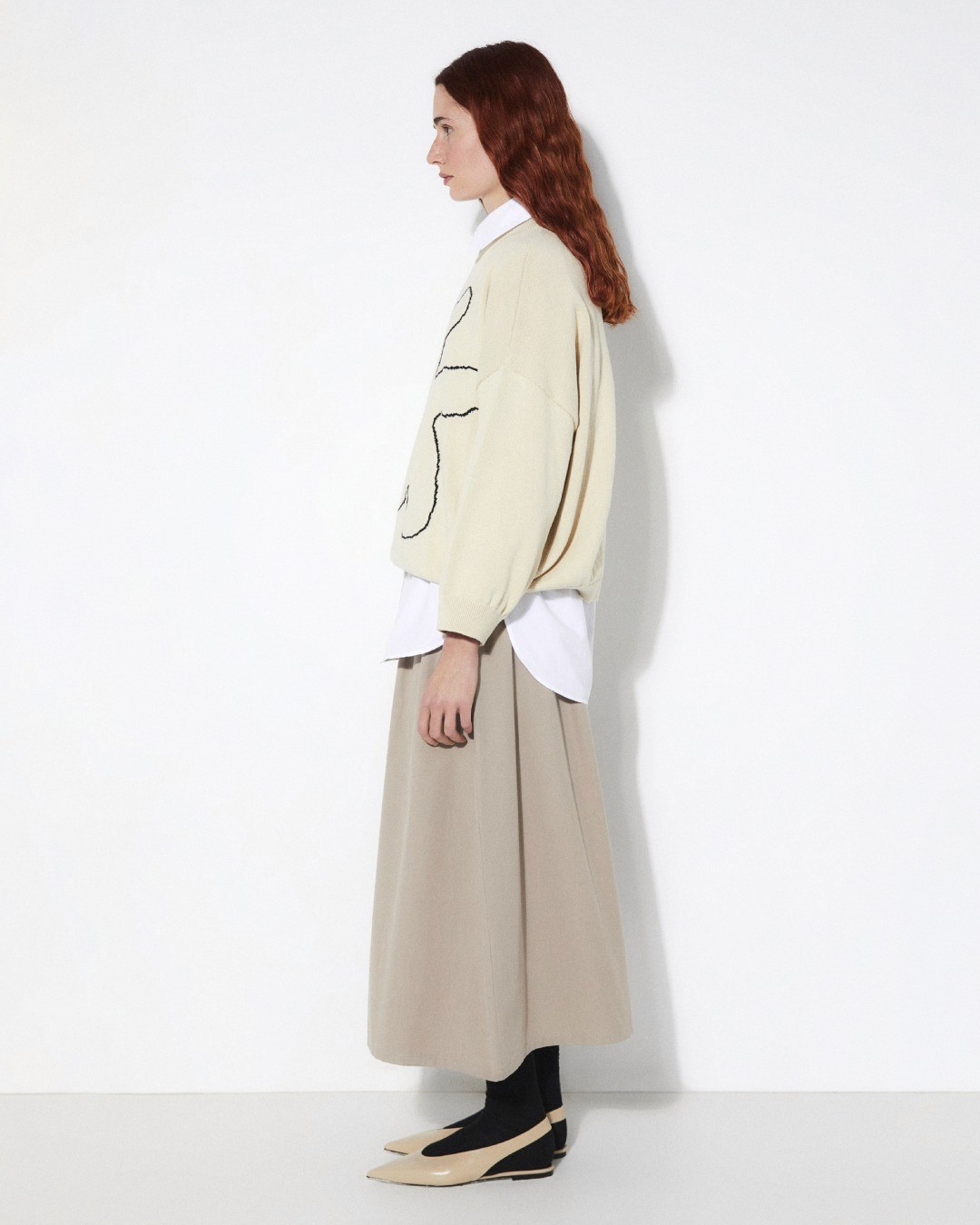 De profil dans le lookbook Rita Row, une mannequin porte une pull blanc cassé, un chemisier blanc dépasse et une jupe longue beige
