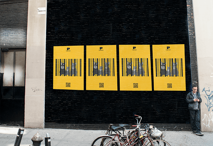 A man stands on the sidewalk next to a black brick wall displaying four identical yellow posters. Each poster features a black barcode with the letters “P” and “P” at the top, followed by four black cat silhouettes. The man wears a dark jacket and dark pants, looking down at his phone or tablet. Bicycles are parked on the sidewalk near the man, and a fire hydrant is visible to the left. The wall shows some graffiti near the right edge.
