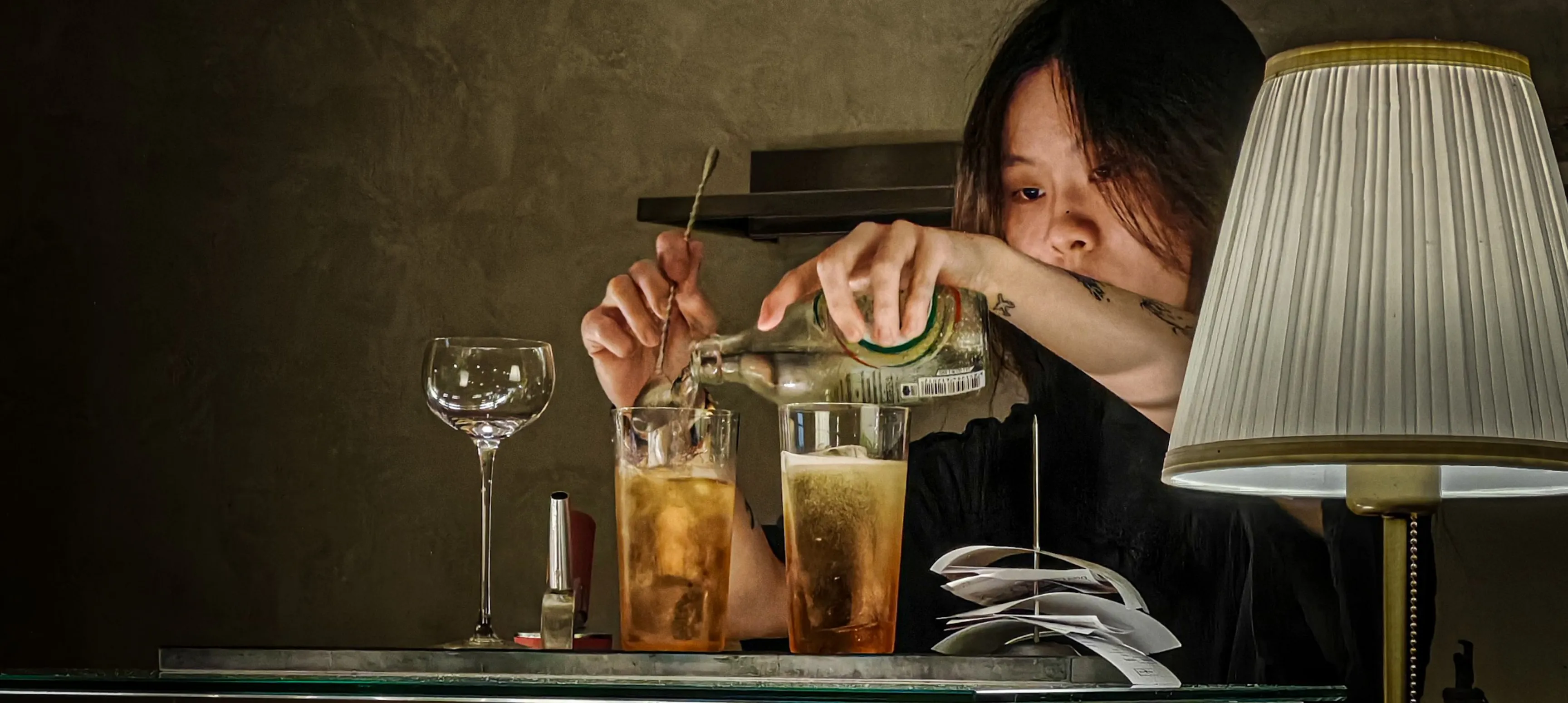 A bartender making cocktails in a dimly lit bar with a lamp beside them. 
