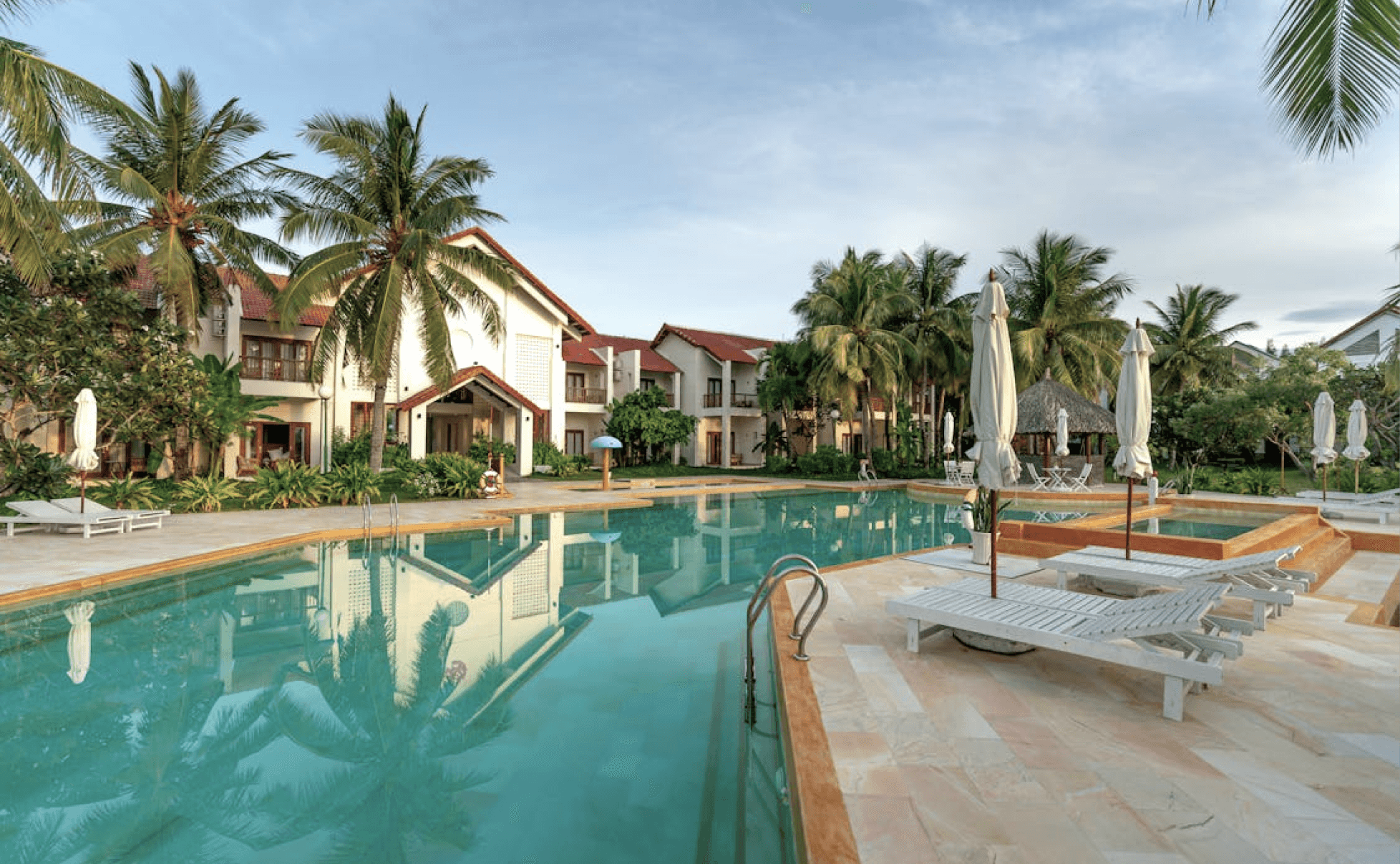 Luxury beach resort with white modern buildings, pergolas, lounge seating, greenery, and rectangular pool on tiled patio.

