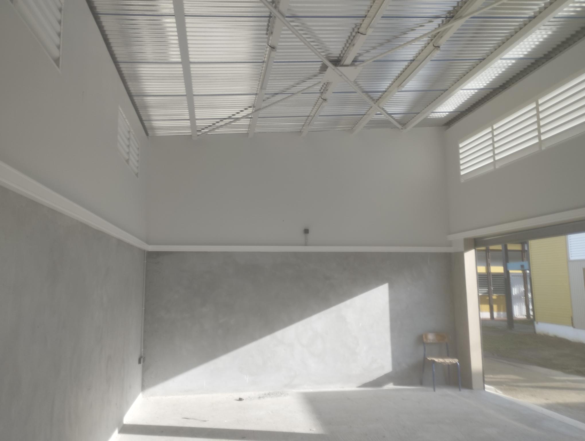 Une pièce ensoleillée et vide, avec des murs gris, un toit métallique et des fenêtres à ventilation. Une chaise en bois se trouve seule dans le coin arrière, projetant de longues ombres.