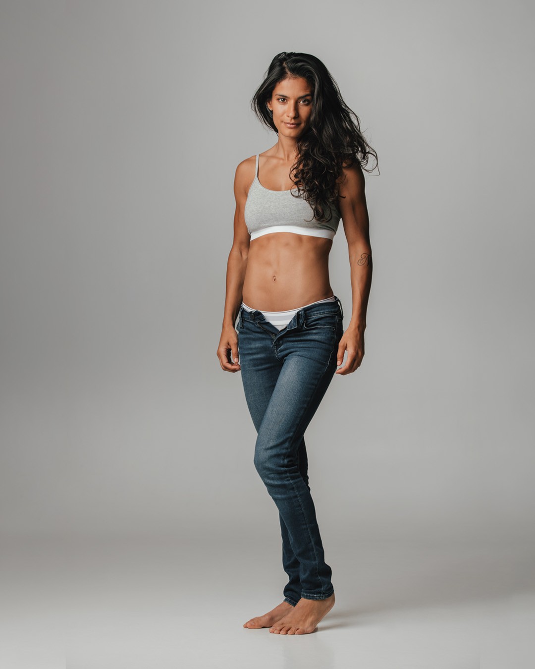 Photo de studio en pleine longueur d'une femme à la peau mate et aux longs cheveux ondulés bruns et foncés. Elle est debout, pieds nus, portant un soutien-gorge de sport gris clair et un jean délavé, déboutonné à la taille. Son expression est neutre ou légèrement souriante, son corps est athlétique. L'arrière-plan est un fond uni gris clair.