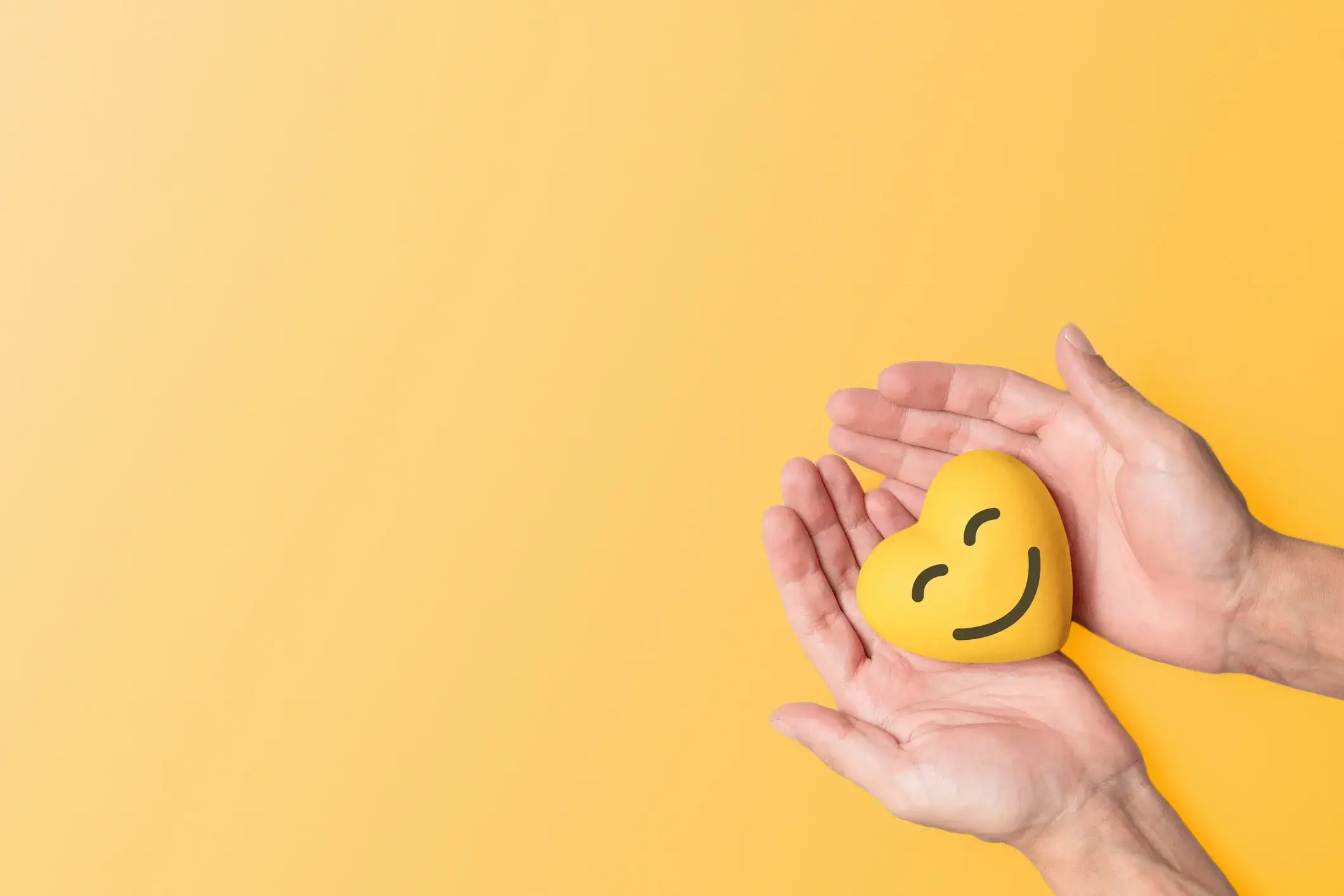 Two hands gently holding a yellow smiley face heart, symbolising care and support