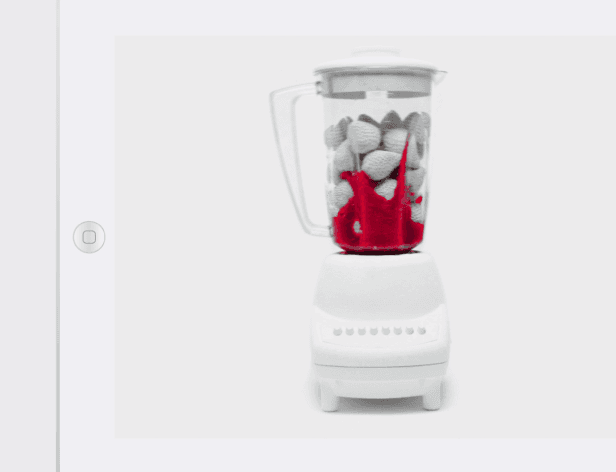 A white blender with a clear glass jar filled with white, rounded objects sits on a white surface. A red liquid is visible inside the jar. The blender has a white base with visible buttons and controls.