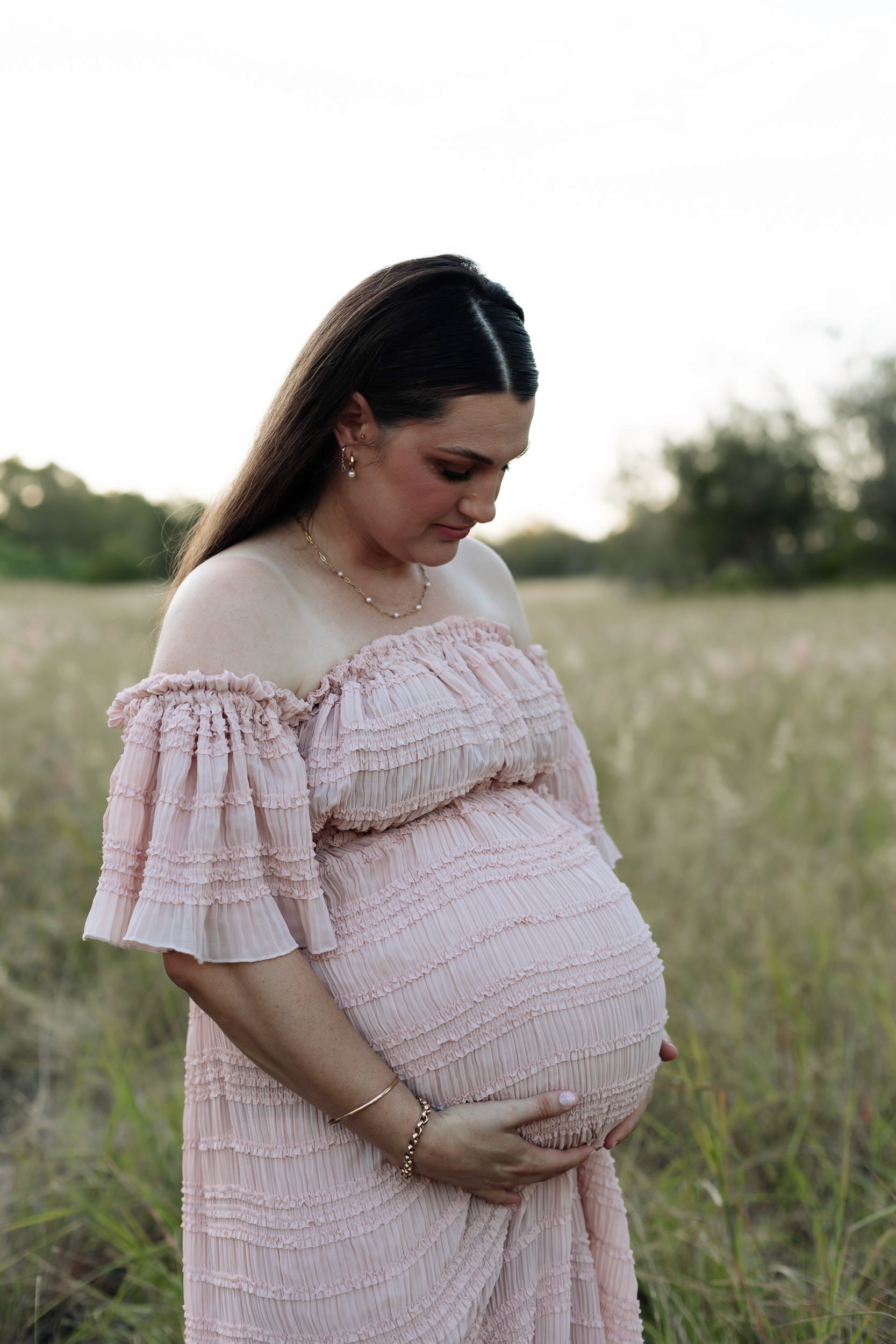 Expecting mother walking through sunlit long grass at sunset captured during outdoor maternity session in Mackay
