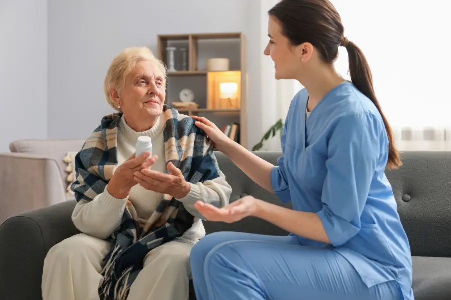 Home care nurse in blue scrubs reviewing medication with an elderly woman wrapped in a plaid blanket on a couch.