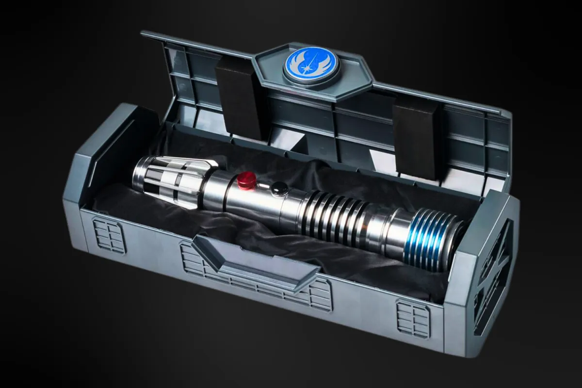 A high-quality replica of Plo Koon’s Legacy Lightsaber hilt displayed inside a sleek, black storage case with a blue Jedi Order emblem on the lid. The hilt features a polished silver design with blue and black accents, along with red and black activation buttons. The case is lined with soft black fabric, enhancing the collectible presentation.