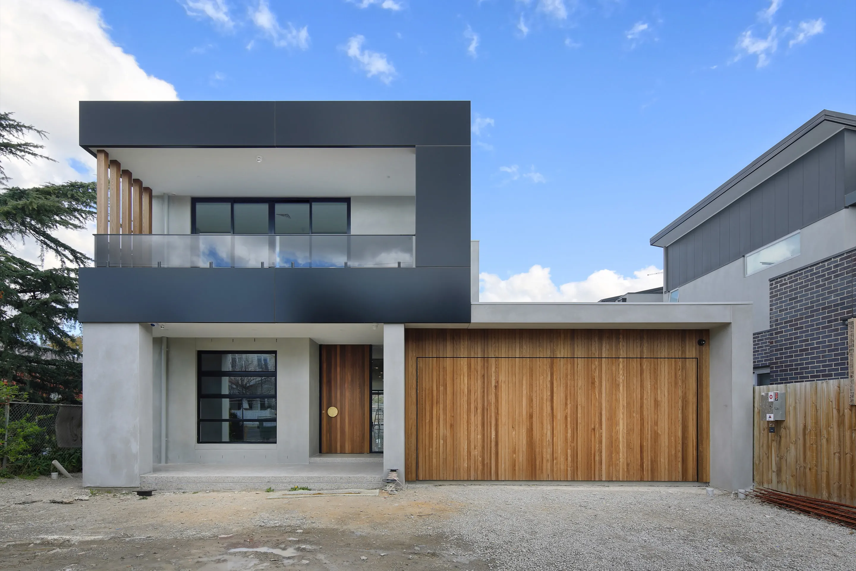 Front of the contemporary Coburg home, a two-storey residence with a charcoal-clad cantilevered upper box on timber posts, rendered ground floor, black-framed windows, a timber pivot door and a timber-clad double garage