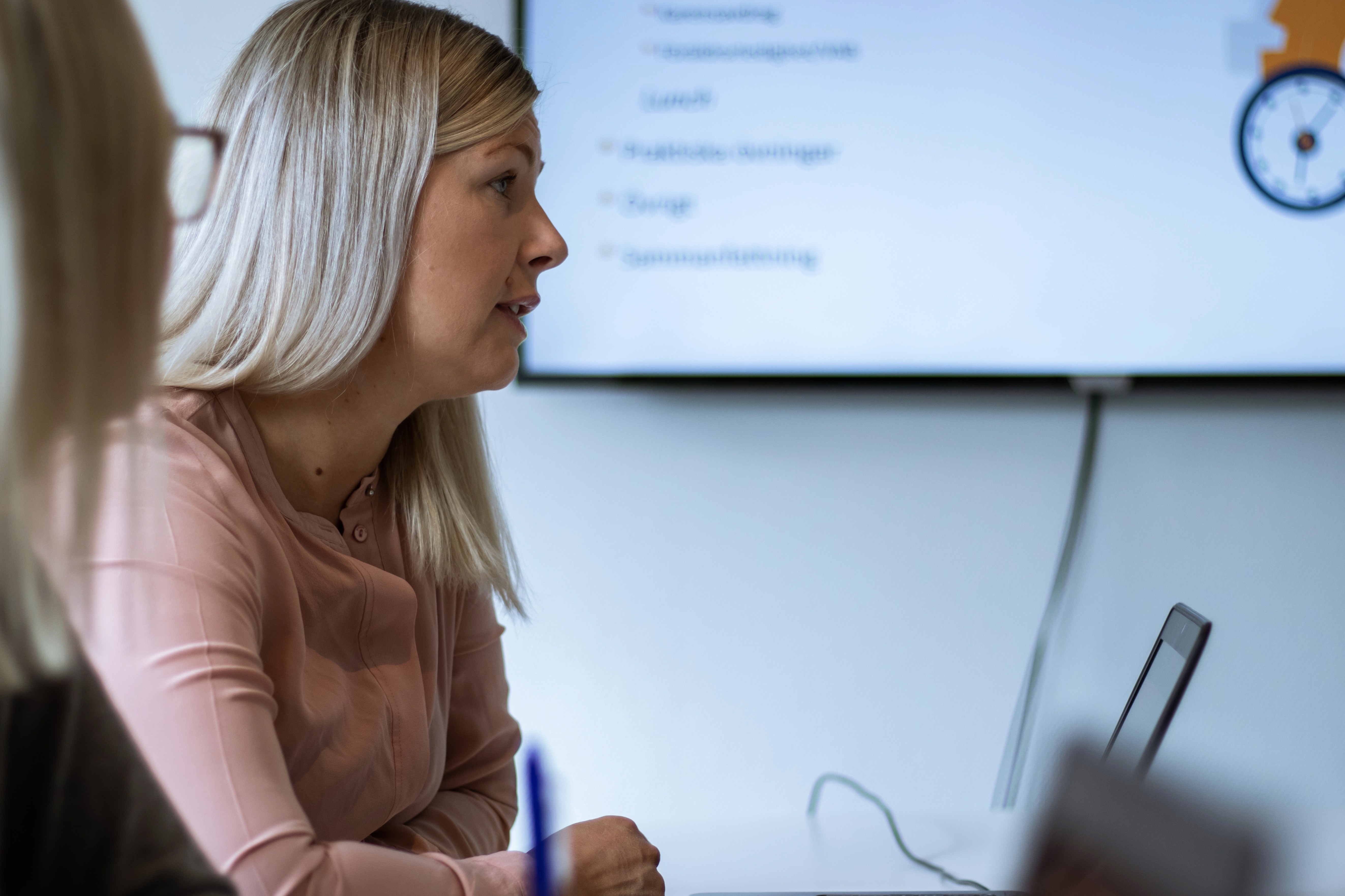 Kvinna i rosa blus talar på möte, tittar åt sidan. Laptop på bordet.