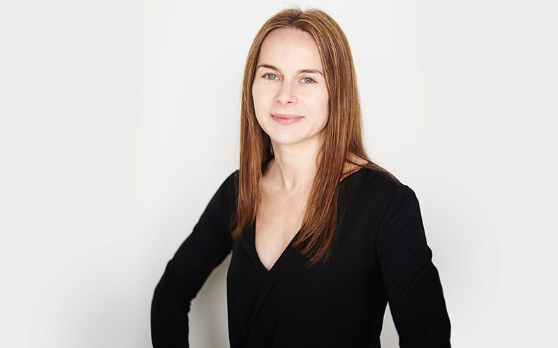 A photo of a woman wearing a black blouse and sitting in front of a white wall.