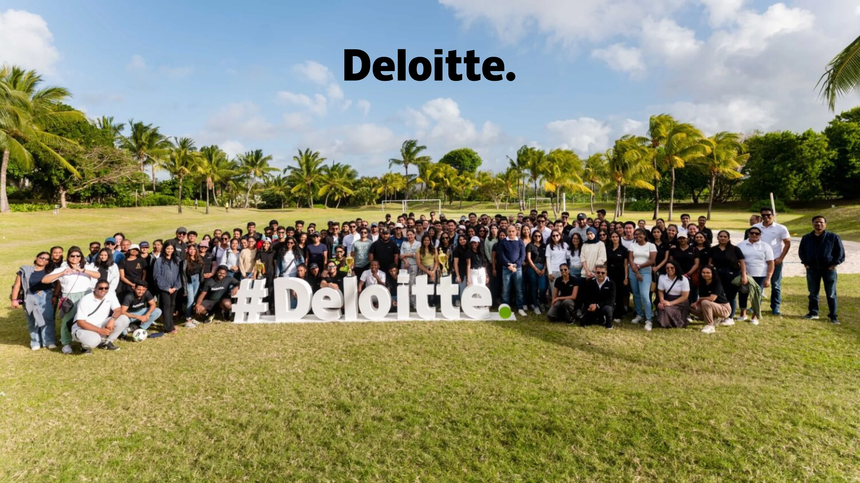 Deloitte large team photo showing over 100 employees gathered outdoors on a lawn in front of palm trees, with a large Deloitte hashtag sign and the Deloitte wordmark displayed