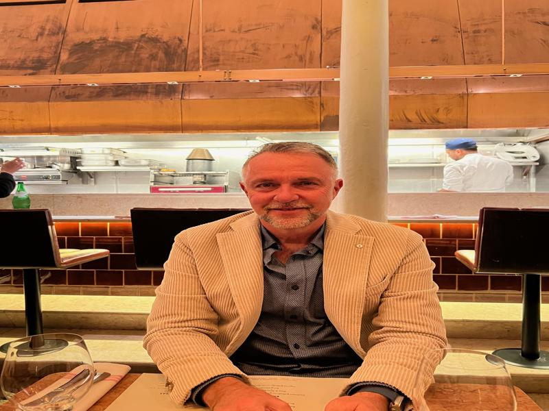 A person sitting at a restaurant table with a view of the open kitchen in the background, wearing a beige striped blazer and a blue shirt, with a chef visible cooking behind them.