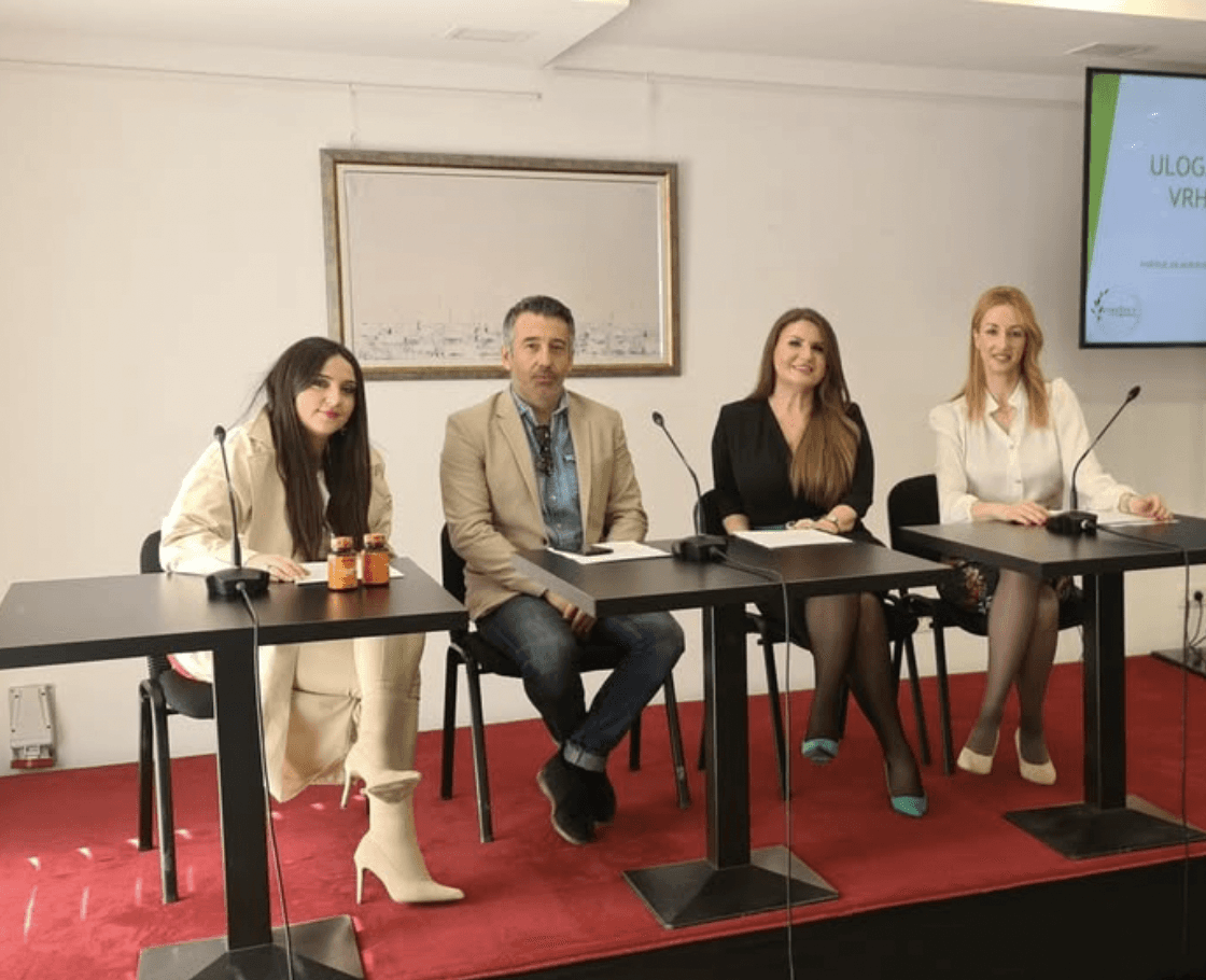A person wearing a beanie and glasses is working at a computer desk in a sunlit apartment, surrounded by plants, with a cityscape visible through the large windows.
