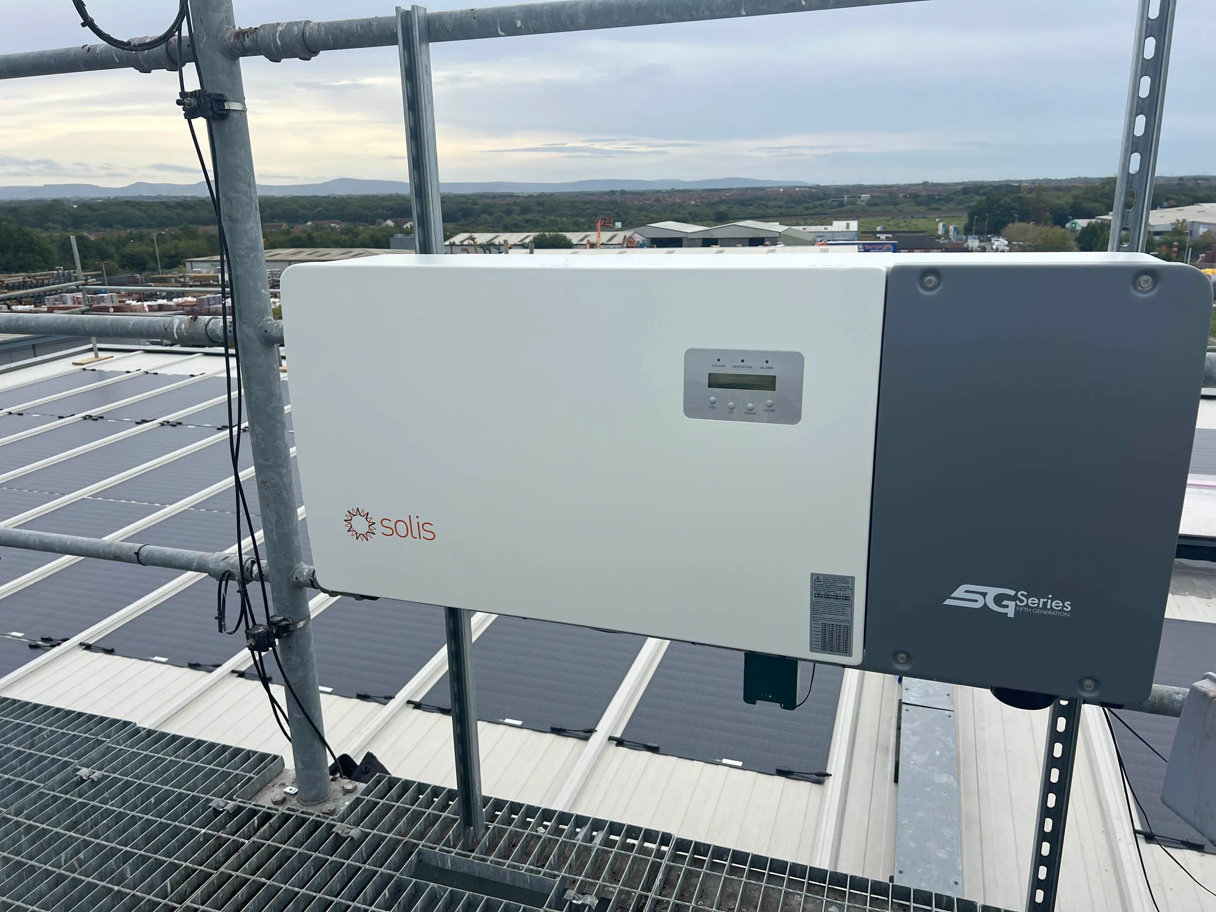 Industrial rooftop with a mounted white Solis SG Series solar inverter, surrounded by solar panels and scaffolding, overlooking a landscape with distant hills and a partly cloudy sky.