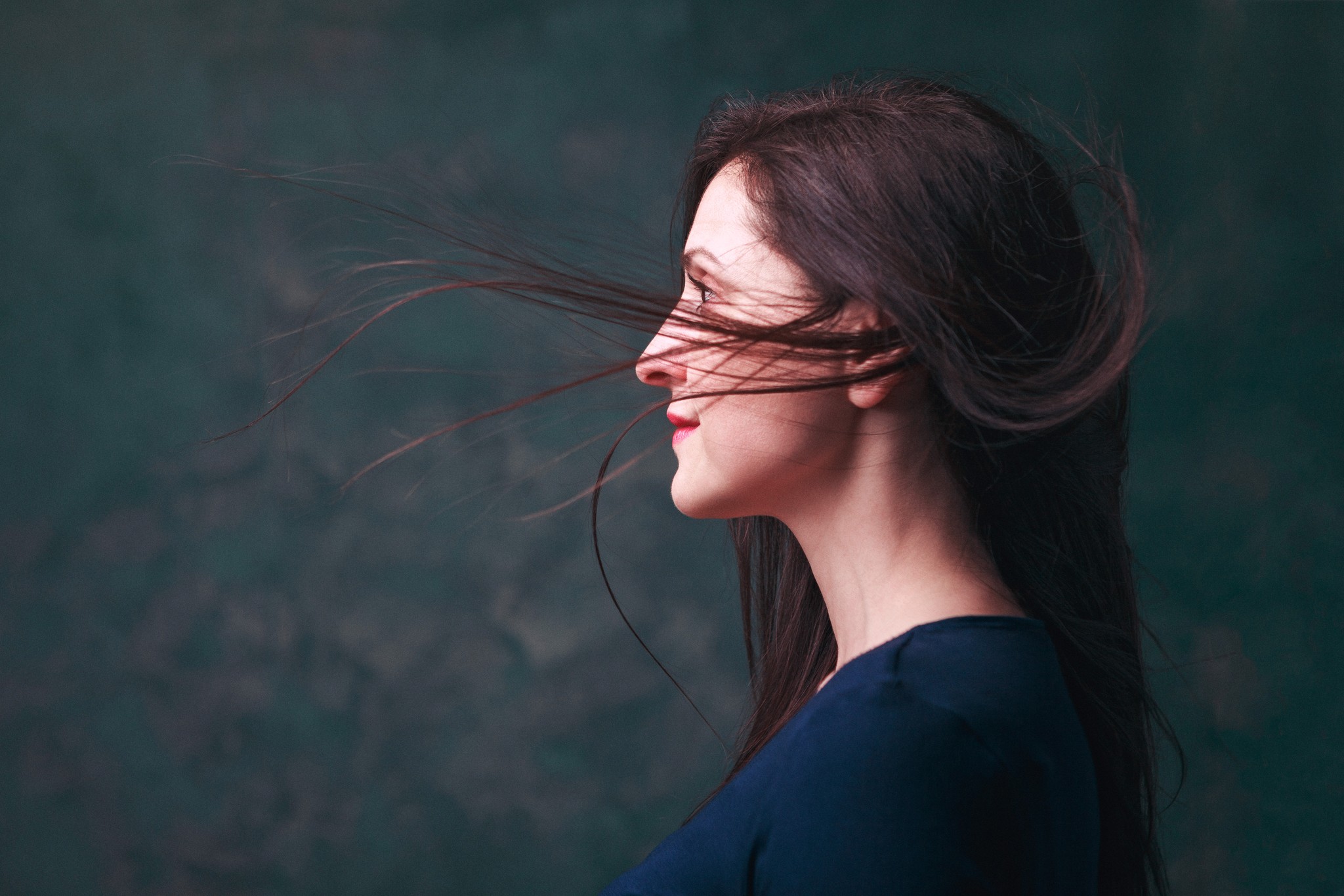 Editorial portrait of an actress in profile with wind-swept hair and soft cinematic lighting against a dark textured backdrop.