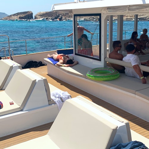 People relaxing on the deck of a boat, some sunbathing while others sit under a shaded area. The sea and rocky coastline are in the background.