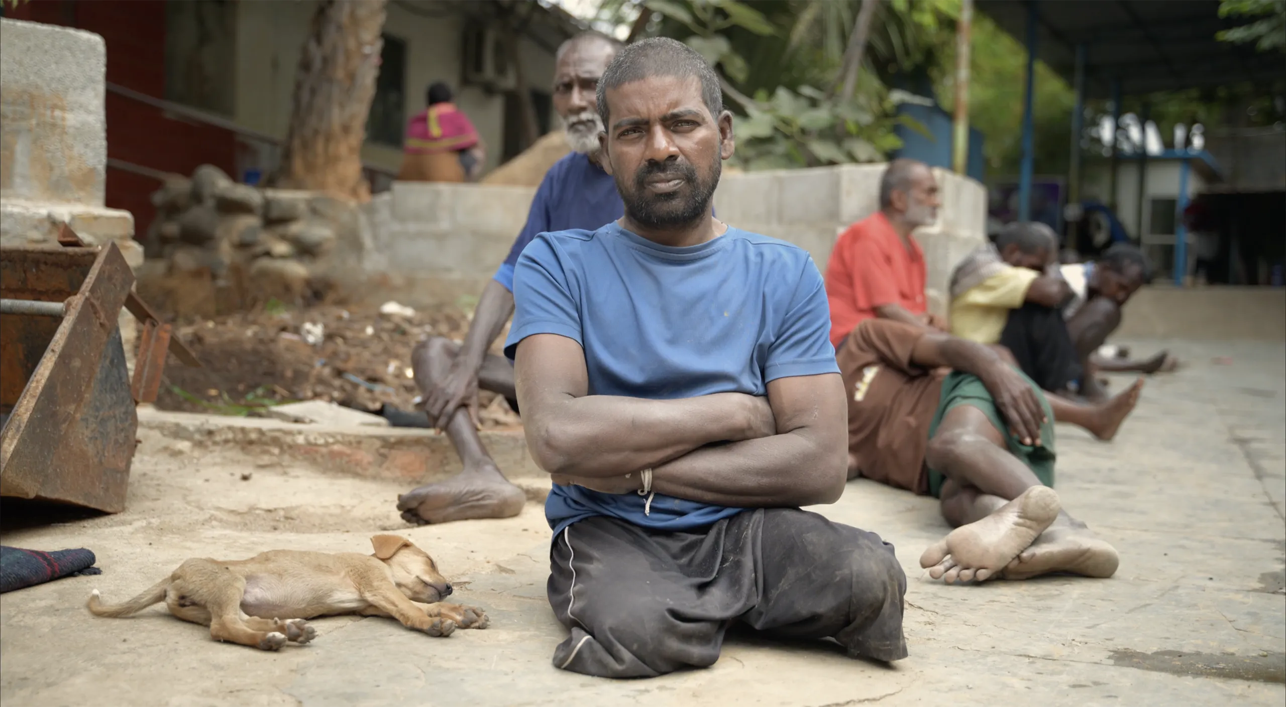 A candid documentary shot of a group of men sitting together on the ground in a village or shelter environment.