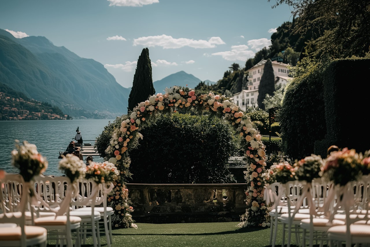 A scenic outdoor wedding venue with mountains, a floral arch, and rows of chairs set by a lake.