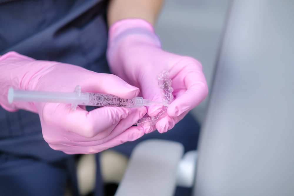 A close-up view of a dental professional's hands wearing pink protective gloves, demonstrating the application of home whitening gel. They are holding a clear, custom-fit whitening tray and carefully injecting gel from a syringe labeled "BOUTIQUE" into the tray.