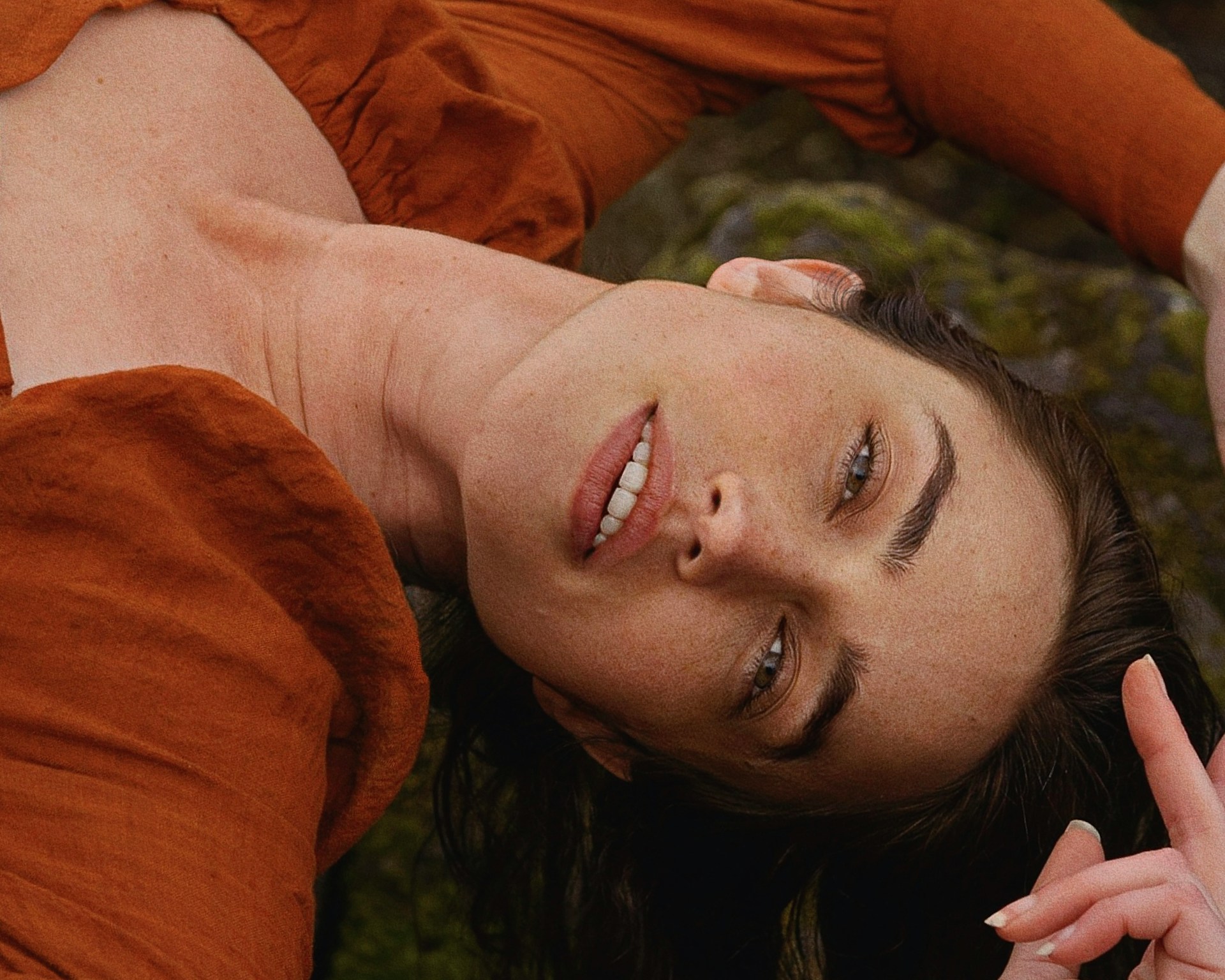 A close-up, tilted portrait of a woman with freckles and brown hair lying down, wearing a rust-colored top.