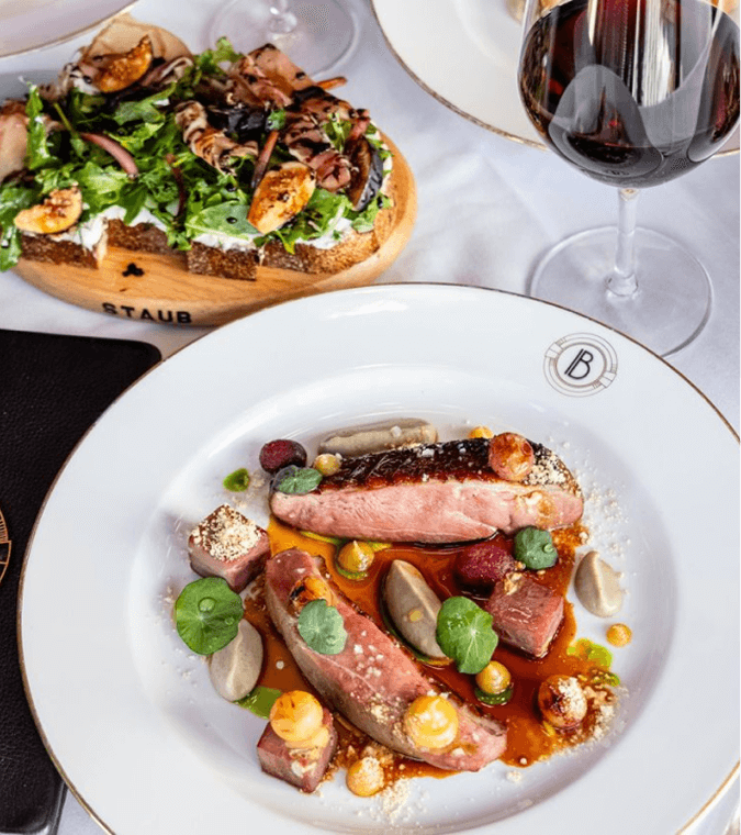 A plated dish of seared duck breast with garnishes, accompanied by a salad-topped toast and a glass of red wine on a white tablecloth.