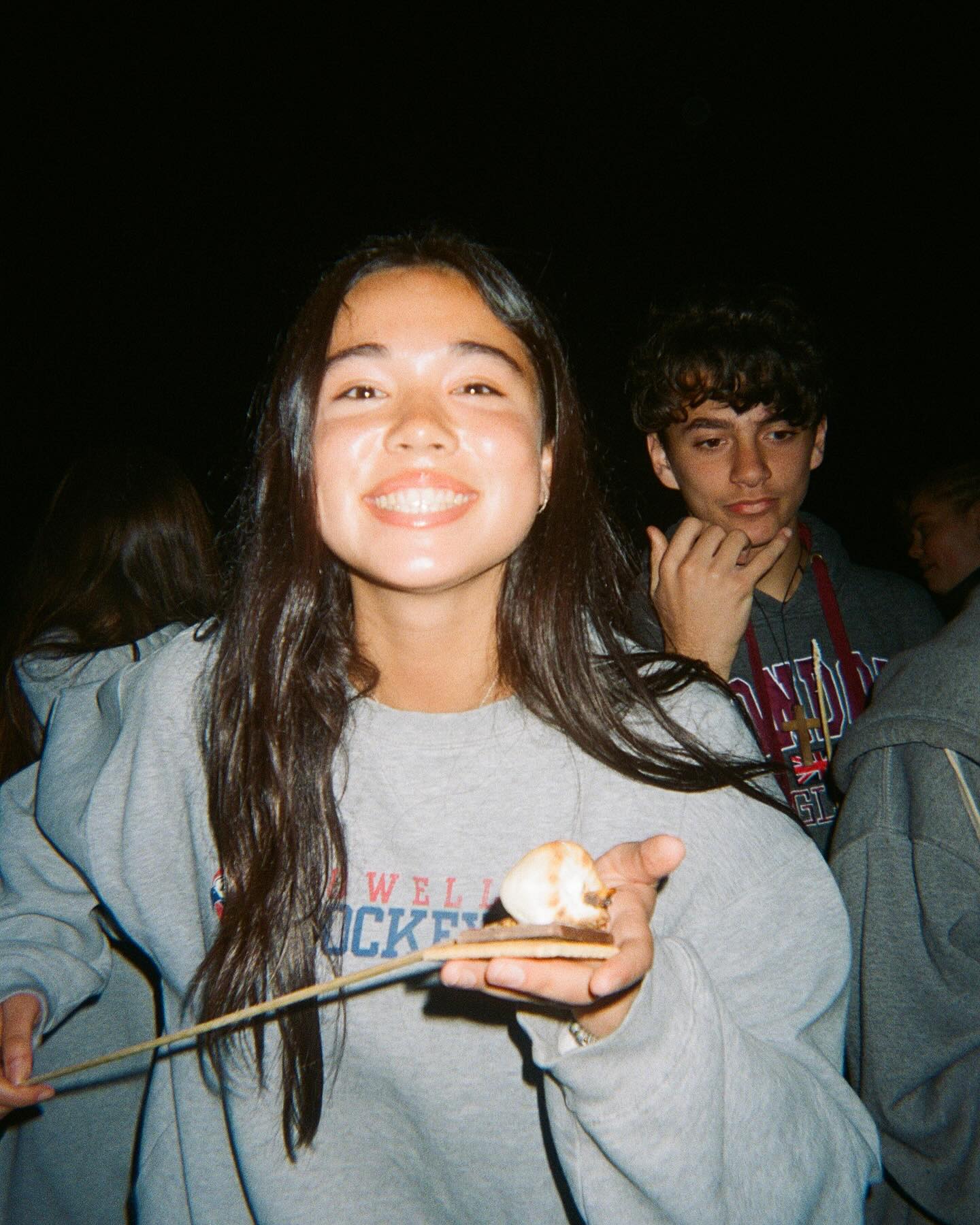 A young person smiling while holding food, with friends behind her.