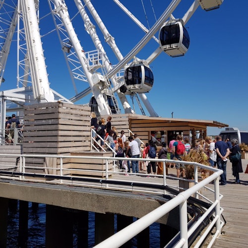 人們在碼頭的摩天輪附近排隊。明亮的藍天和水在背景中。