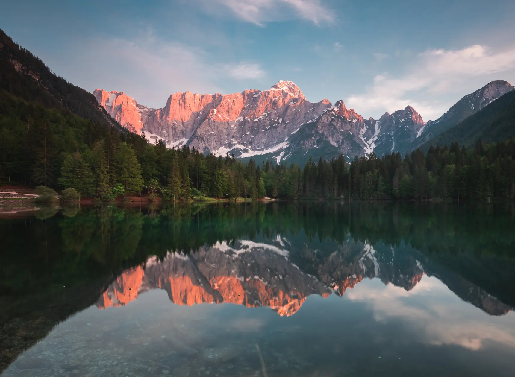 A perfect mirror reflection of the rugged, sunlit Mangart mountains in the calm waters of Lago di Fusine, Italy, surrounded by dense evergreen forests.