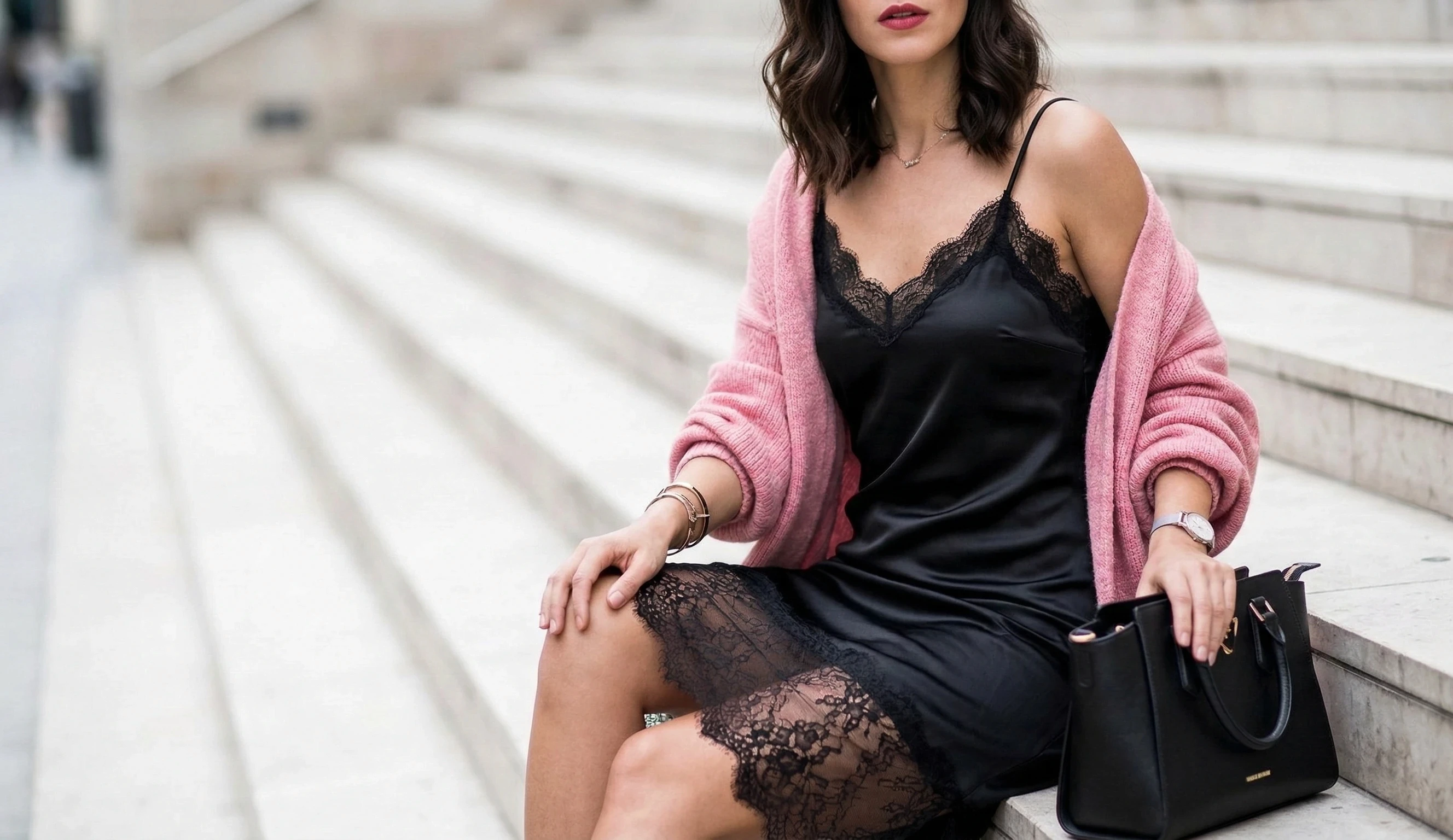 Woman in a black lace slip dress and pink cardigan seated on stone steps.
