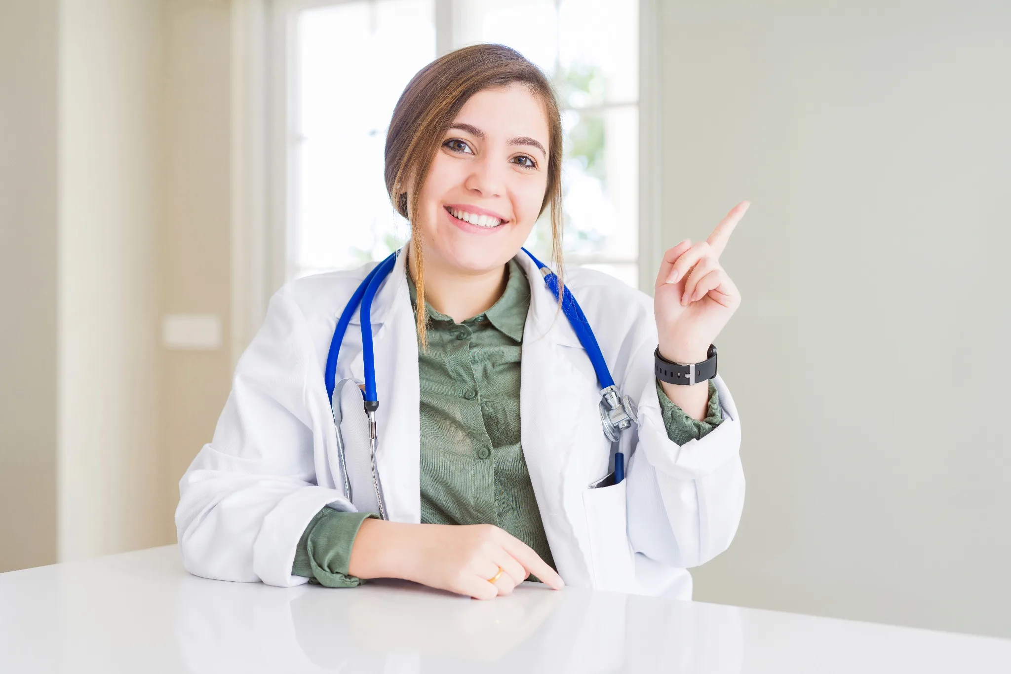 Smiling female physician wearing a white coat with stethoscope around her neck, pointing to the right.